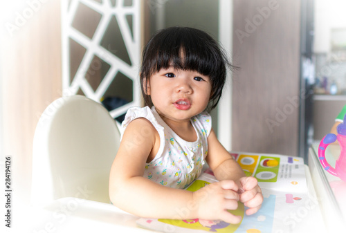 Adorable 18 months baby girl sitting on baby chair and reading book at home