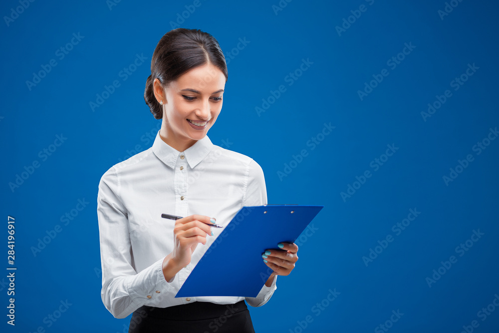 Young businesswoman writing happily on her blue clipboard, isolated on blue background. Young businesswoman writing happily on her blue clipboard, isolated on blue background.