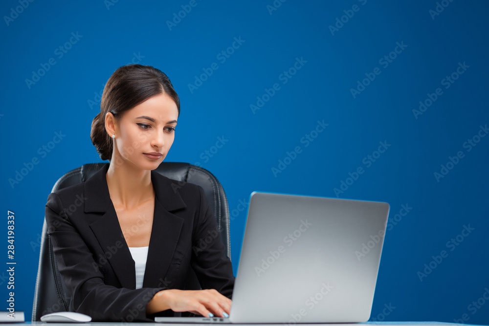 Young businesswoman sitting at her desk focused on her work with the laptop, isolated on blue background.