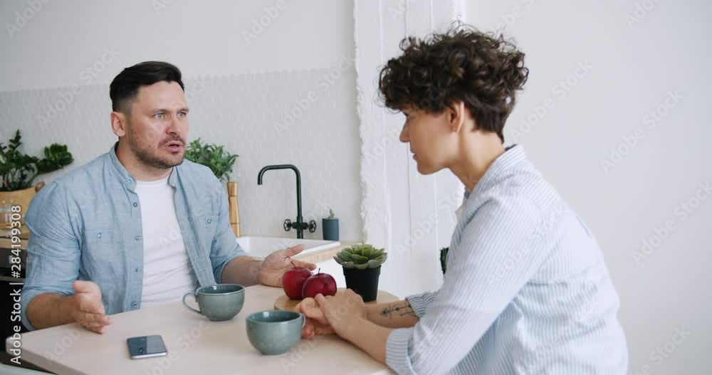 Slow motion of angry man yelling at unhappy wife sitting in kitchen at table fighting discussing problems. Relationship, unhappiness and apartment concept.