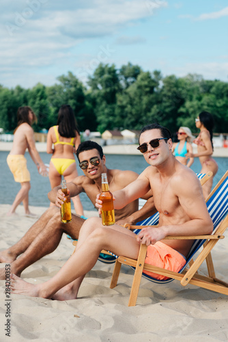 handsome multicultural friends holding bottles of beer and smiling at camera while sitting in chaise lounges on beach © LIGHTFIELD STUDIOS