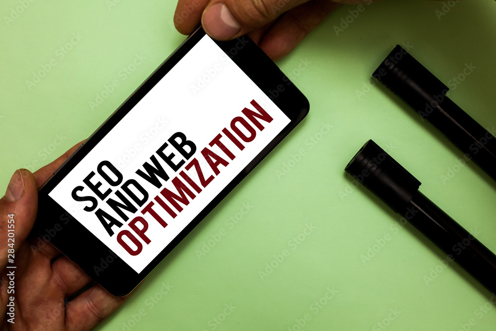 Text sign showing Seo And Web Optimization. Conceptual photo Search Engine Keywording Marketing Strategies Man's hand hold phone with black and red words near two black marker Text sign showing Seo And Web Optimization. Conceptual photo Search Engine Keywording Marketing Strategies Man's hand hold phone with black and red words near two black marker