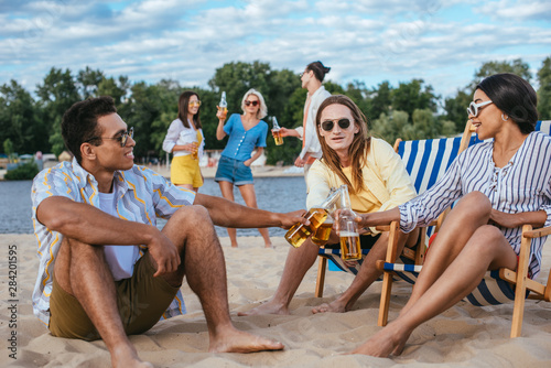 cheerful multiethnic friends clinking bottles of beer while having fun on beach