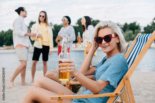 cheerful young woman smiling at camera while sitting in chaise lounge with bottle of beer near multicultural friends