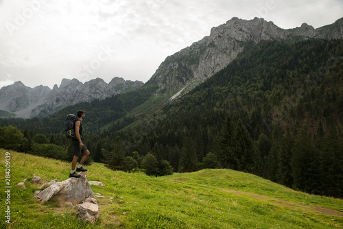 Caucasian man with a trekking backpack standing on a rock and admiring the beautiful mountain landscape. Connection between human being and nature concept