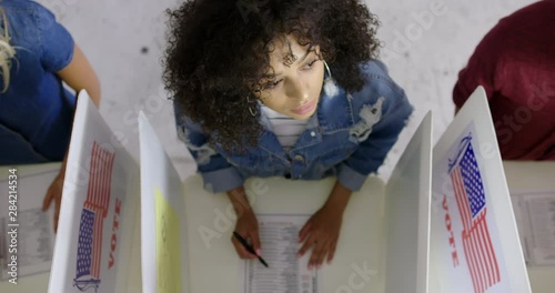 CU overhead view Hispanic/Latina woman looking up and thinking while completing ballots with other young women at voting booths in polling station. Locked off, slow motion 4K 60fps