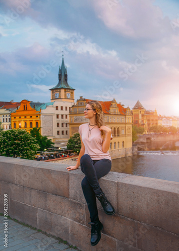 woman sits on a bridge over a river and enjoys the sunset with a smile on her face. Tourist in the old town of Prague. View of multi-colored beautiful houses and buildings. blue jeans, pink t-shirt