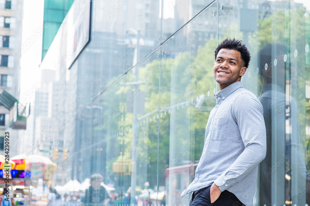 Fototapeta premium Young Mix Race American Man wearing light gray, long sleeve shirt, black pants, hand in pocket, standing against glass wall with reflections on street in Midtown of Manhattan, New York City, smiling..