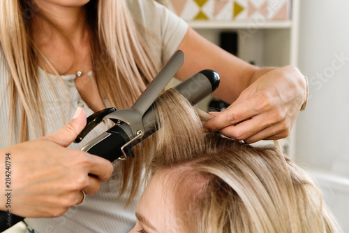 Hairstylist using curling iron of female client in hairdressing salon, closeup view. Hairdressing services. Creating hairstyle. Hair styling process.