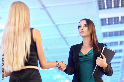 Two young women in the office with folders with papers shake hands