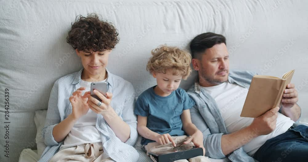 Mother is using smartphone son is holding tablet touching screen while father is reading book in bed at home. Modern family, devices and people concept. Mother is using smartphone son is holding tablet touching screen while father is reading book in bed at home. Modern family, devices and people concept.