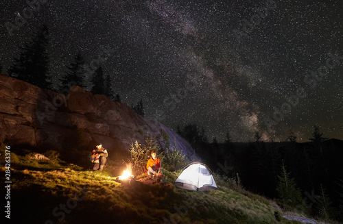 Night camping in the mountains. Happy couple travellers sitting together beside campfire and glowing tourist tent. On background big boulder, forest and night starry sky full of stars and Milky way.