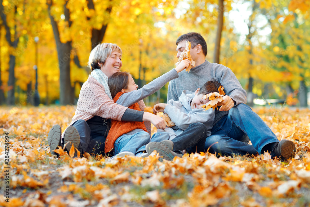 Happy family sitting on fallen leaves, playing and having fun in autumn city park. Children and parents together having a nice day. Bright sunlight and yellow leaves on trees, fall season. Happy family sitting on fallen leaves, playing and having fun in autumn city park. Children and parents together having a nice day. Bright sunlight and yellow leaves on trees, fall season.