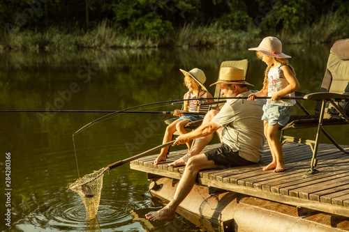 Cute little girls and their granddad are on fishing at the lake or river. Resting on pier near by water and forest in sunset time of summer day. Concept of family, recreation, childhood, nature. photo