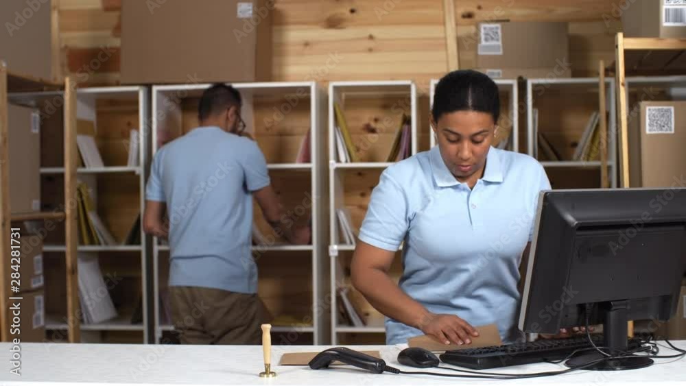 Waist-up shot of black female post office clerk working on shift together with Arab male trainee, and registering fresh mail on computer and instructing while he is sorting letters into compartments