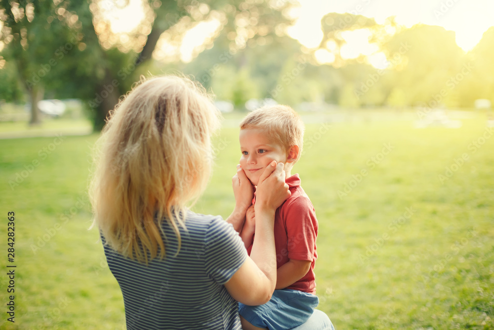 Young Caucasian mother pulling boy toddler cheeks. Mom sitting together with son on summer day in park and showing her love and affection. Happy family childhood lifestyle. Cute authentic moment. Young Caucasian mother pulling boy toddler cheeks. Mom sitting together with son on summer day in park and showing her love and affection. Happy family childhood lifestyle. Cute authentic moment.