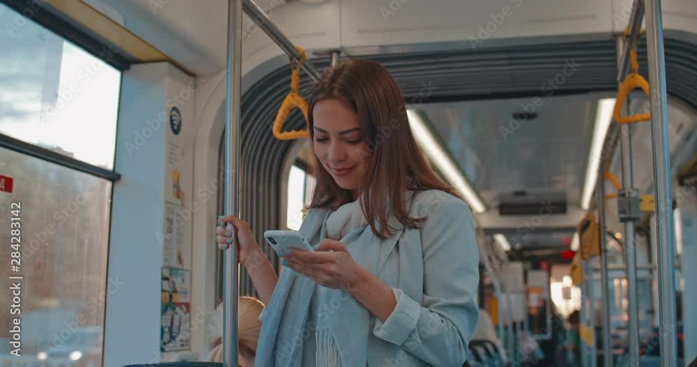 Portrait of cute girl holds the handrail, texting and browsing on mobile phone in public transport. Smiling woman using the smartphone. City, urban background Portrait of cute girl holds the handrail, texting and browsing on mobile phone in public transport. Smiling woman using the smartphone. City, urban background