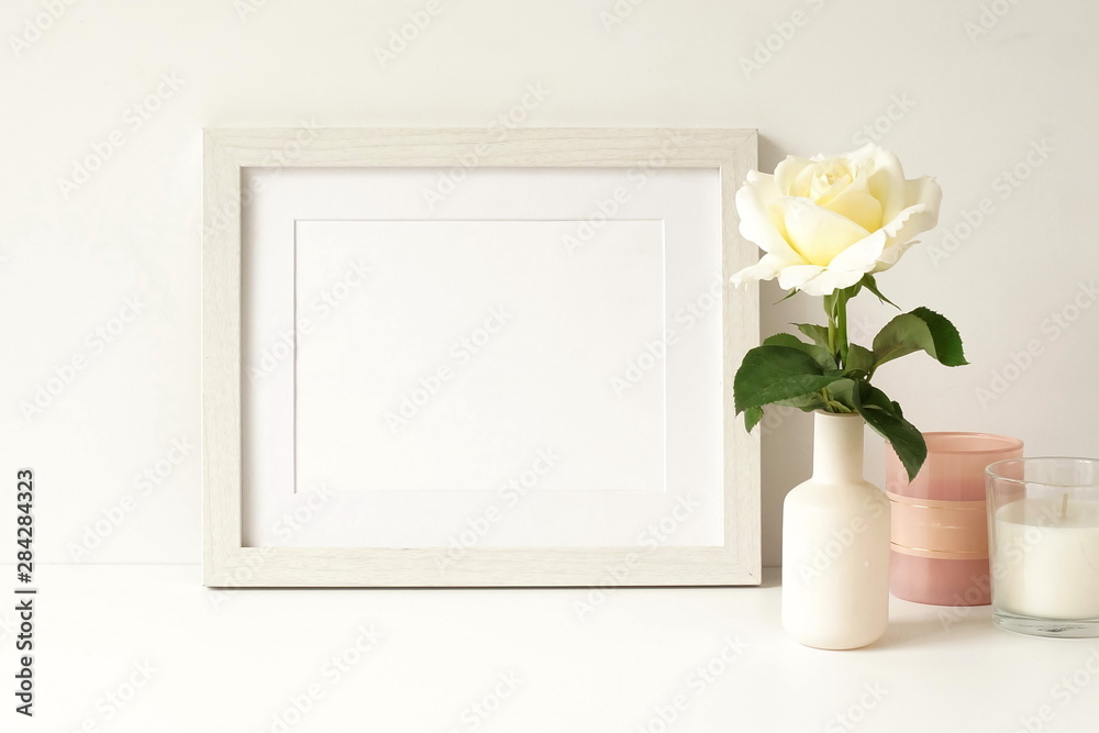 White frame mock up on white wall, rose in a vase, decorative candles . Home decor close up. Copy space