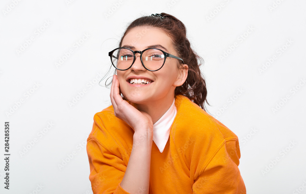 Portarit of cheerful student girl smiling broadly, wears orange sweater and round transparent eyewear. Pretty positive young woman feels joyful posing over white studio wall. People and emotions Portarit of cheerful student girl smiling broadly, wears orange sweater and round transparent eyewear. Pretty positive young woman feels joyful posing over white studio wall. People and emotions