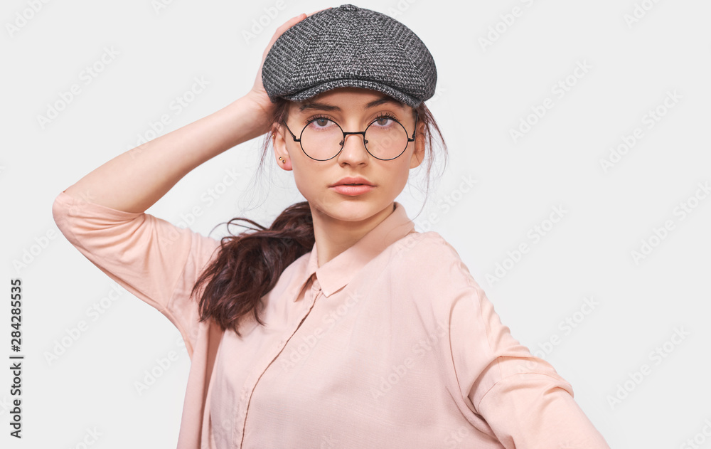 Beautiful young woman wears transparent round spectacles, casual pink shirt and gray cap, looks seriously directly into camera, poses in studio. People emotions concept Beautiful young woman wears transparent round spectacles, casual pink shirt and gray cap, looks seriously directly into camera, poses in studio. People emotions concept
