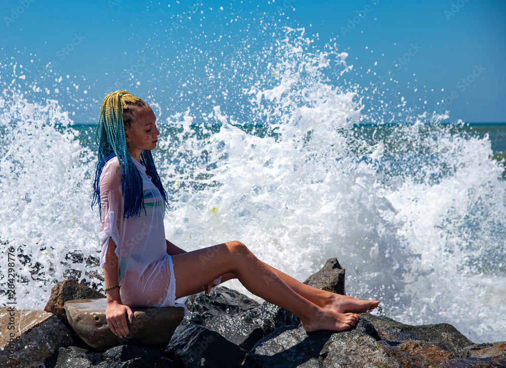 A girl with blue Senegalese pigtails is sitting on the stones. Waves are breaking on the stones and splashes are scattered in different directions. Sunny, bright, funny photo.