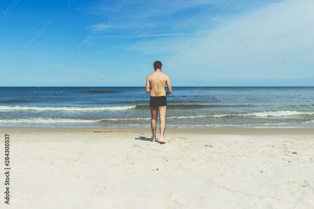 Young guy walking on an empty sandy beach towards the sea - silhouette of a young unrecognizable man shot from behind Young guy walking on an empty sandy beach towards the sea - silhouette of a young unrecognizable man shot from behind