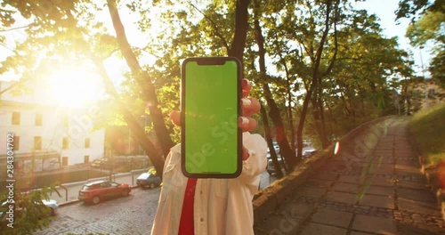 Portrait of woman showing vertical mobile mockup greenscreen phone outdoor. Close-up girl tourist holding smartphone on camera standing at sunset in citu center. photo