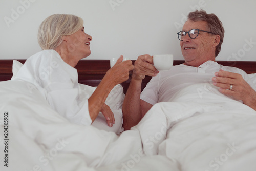 Active senior couple having coffee together in bed in bedroom at comfortable home