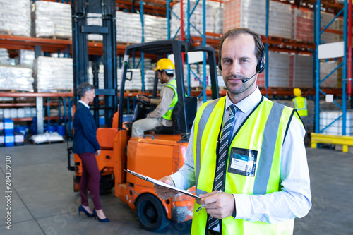 Male supervisor with headset and clipboard looking at camera in warehouse