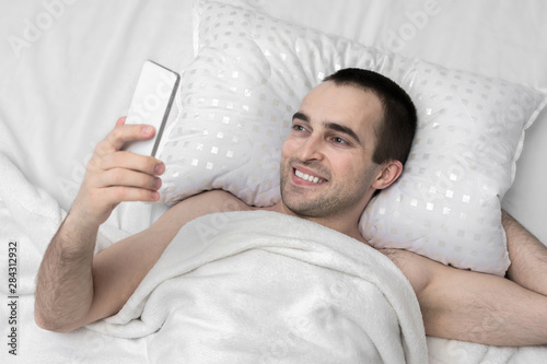 Joyful man woke up in the morning in his bedroom, man uses smart phone, top view