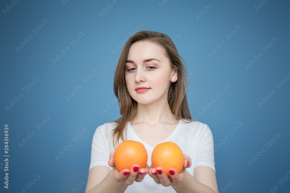 Beautiful girl smiling and offer you an oranges at the camera, looking at oranges, blue background, copy space Beautiful girl smiling and offer you an oranges at the camera, looking at oranges, blue background, copy space