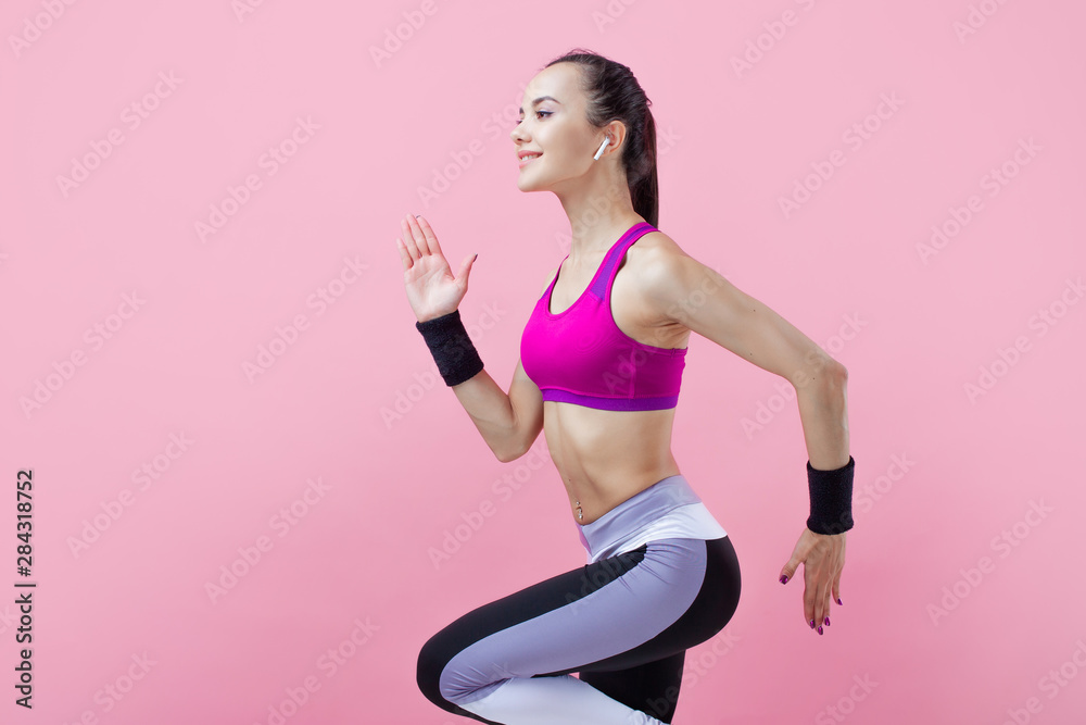 A young attractive girl runs with smile, wireless headphones and in a bright sports top, on a pink background, cardio.