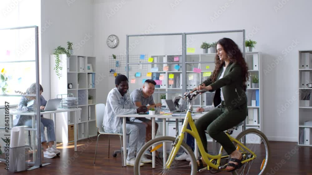 Slow motion shot of young beautiful woman riding bike in open space office while group of cheerful multiethnic colleagues smiling and looking at her Slow motion shot of young beautiful woman riding bike in open space office while group of cheerful multiethnic colleagues smiling and looking at her