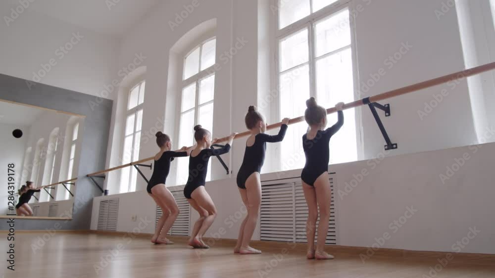Back view of four little Caucasian girls in black costumes standing in row and jumping using stick on the wall
