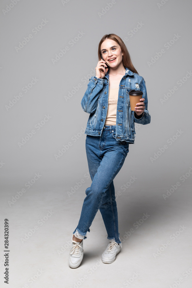 Young woman talking by mobile phone drinking coffee posing isolated over grey wall background Young woman talking by mobile phone drinking coffee posing isolated over grey wall background