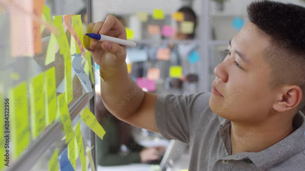 Close up view of young Asian man writing business tasks on colorful sticky notes attached to glass wall while working in the office