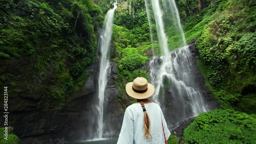 Back of woman in straw hat is enjoying incredible high waterfall Sekumpul (95m) in Bali rainforest. Beautiful wild nature of Indonesia photo