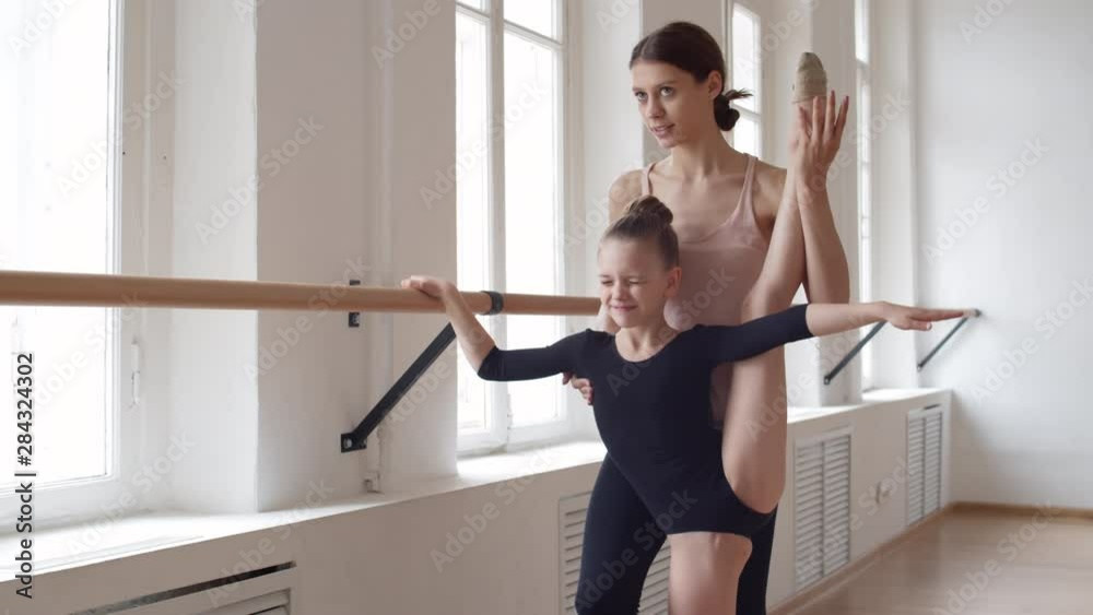 Medium shot of young female ballet teacher helping little female ballerina in black costume standing at barre doing vertical twine