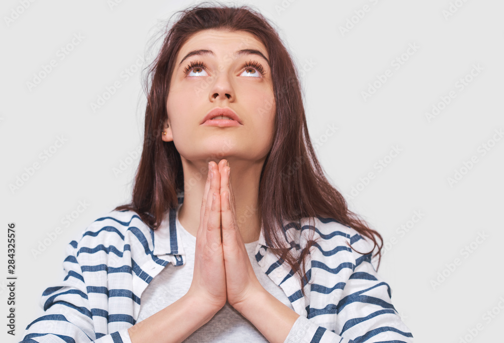 Candid shot of young woman praying for her family, looking up. Meditating female wearing striped shirt holding hands together in praying gesture, feeling thankful. People and body language concept Candid shot of young woman praying for her family, looking up. Meditating female wearing striped shirt holding hands together in praying gesture, feeling thankful. People and body language concept