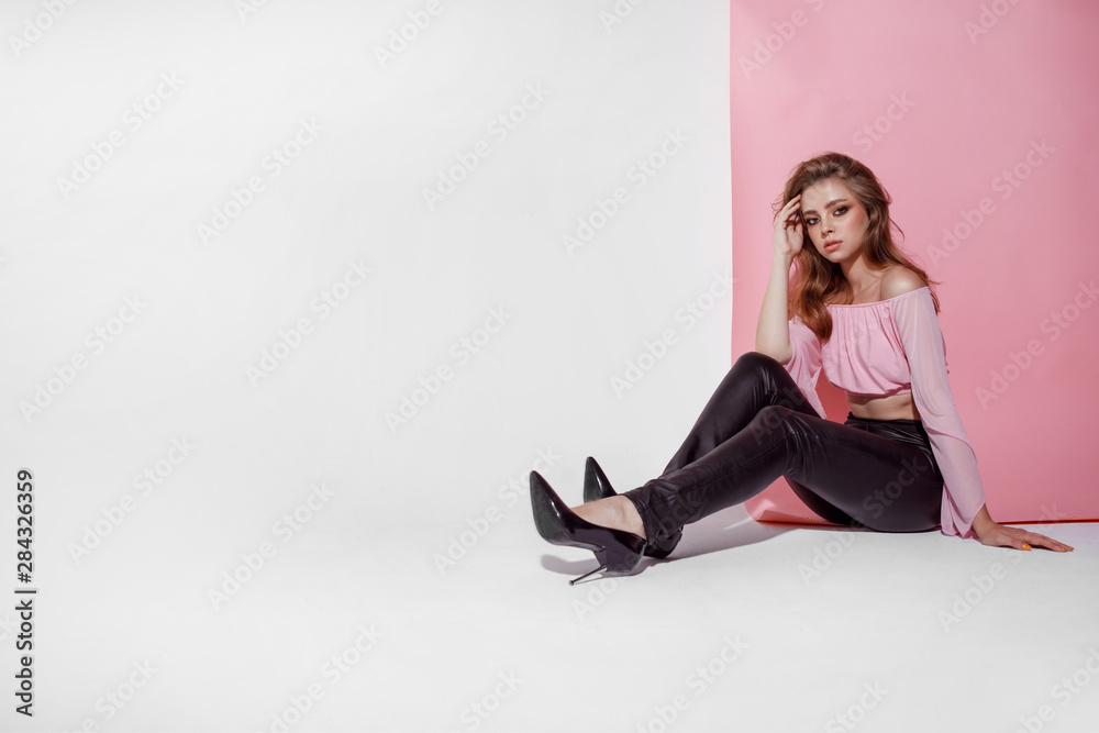 A lovely young female with professional make-up sits like a doll with long legs on a pink white background in a short top and leather leggings, copyspase A lovely young female with professional make-up sits like a doll with long legs on a pink white background in a short top and leather leggings, copyspase