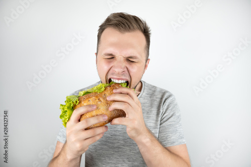young handsome hungry man with closed eyes biting fresh sandwich with salad leaf on isolated white background