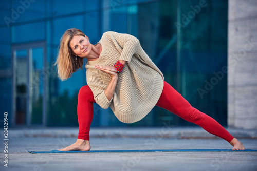 Beautiful young woman practices yoga asana Parivritta Parshvakonasana - Revolving side angle posture outdoors against the background of a modern city photo