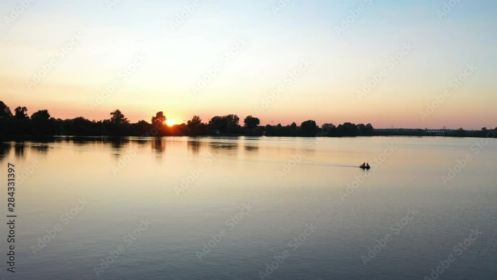 Picturesque Lake Sunset Landscape Aerial. Silhouettes of Couple in Boat on Water and Grove on Coast Picturesque Lake Sunset Landscape Aerial. Silhouettes of Couple in Boat on Water and Grove on Coast