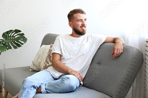 Bearded guy wearing blank white t-shirt & denim pants sitting alone at home on grey textile couch. Young man w/ facial hair in domestic situations. Interior background, copy space, close up, monstera.