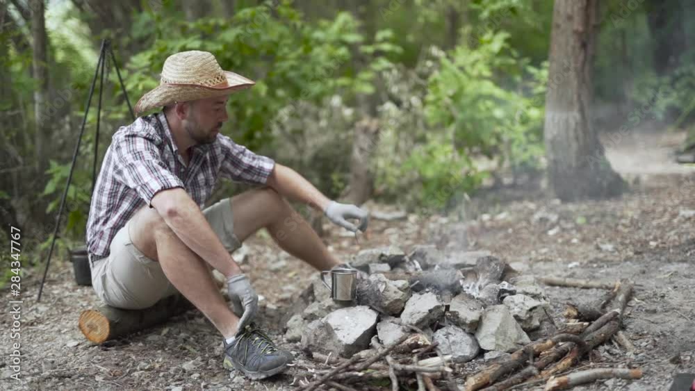 A man in a straw hat adding wood to a bonfire and picking up a tin cup from the side A man in a straw hat adding wood to a bonfire and picking up a tin cup from the side