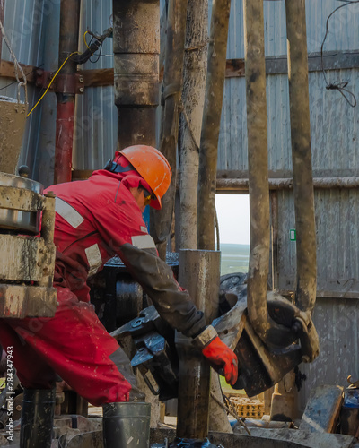 Work driller in red uniform, in helmet and goggles. He with the help of an elevator hangs drill pipes to lift them from an oil well and continue its drilling. The concept of a working person.