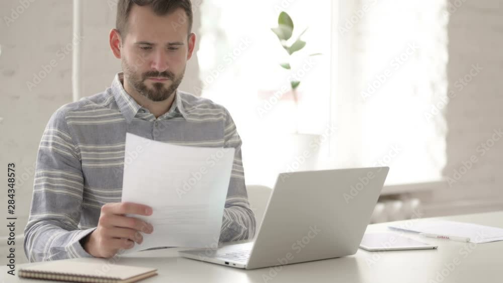 Casual Young Man Doing Paperwork and Typing on Laptop Casual Young Man Doing Paperwork and Typing on Laptop