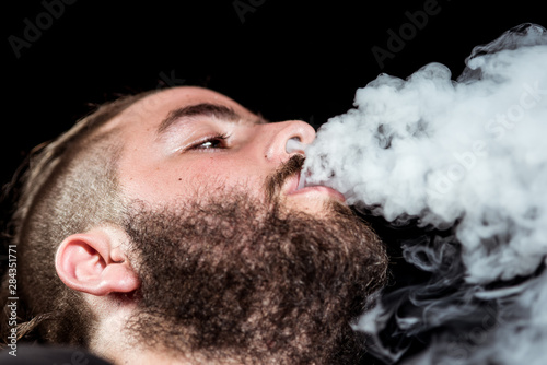 Young man with a leafy beard exhales smoke through his mouth and nose on black background