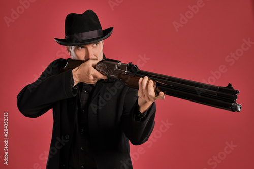 Middle-aged man with beard, mustache, in black jacket and hat, holding a gun while posing against a red background. Sincere emotions concept. photo