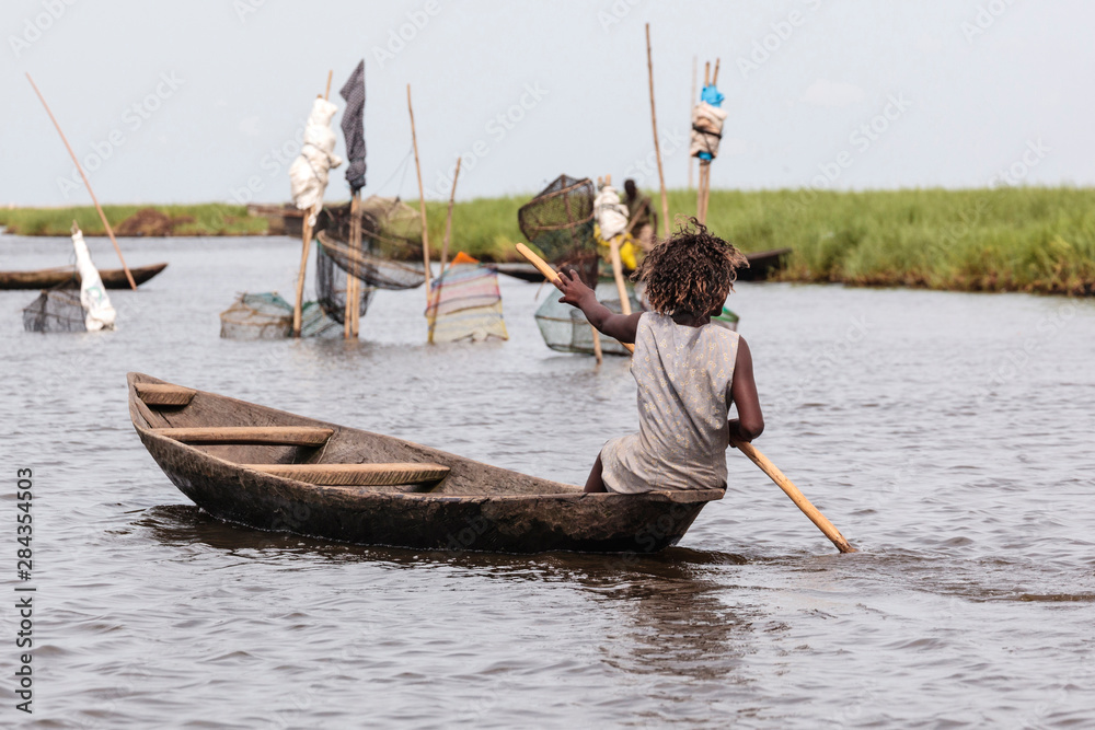 Africa, Benin, Ganvie. Young woman paddling dugout canoe with fishing nets in background.
